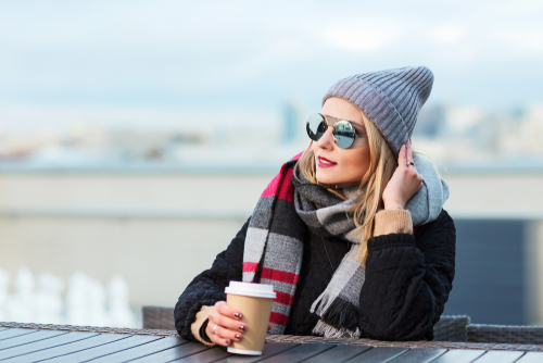 woman in winter clothing and sunglasses sitting at outdoor table while holding cup of coffee